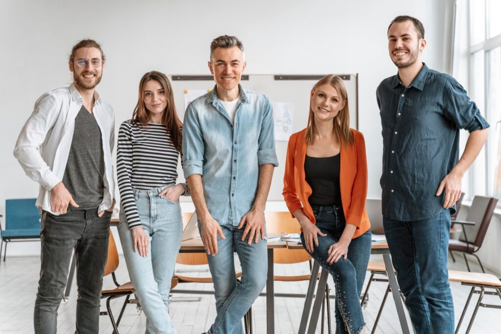 Fem leende studenter står på rad framför bord och stolar i ett klassrum.
